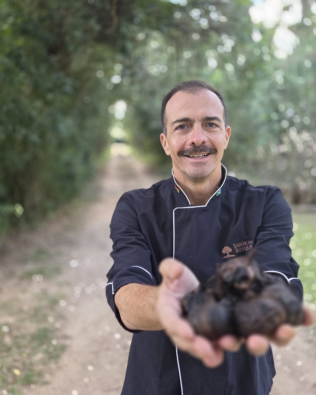O produtor da Sabor do Bosque segurando bulbos de alho negro em um caminho de terra entre árvores