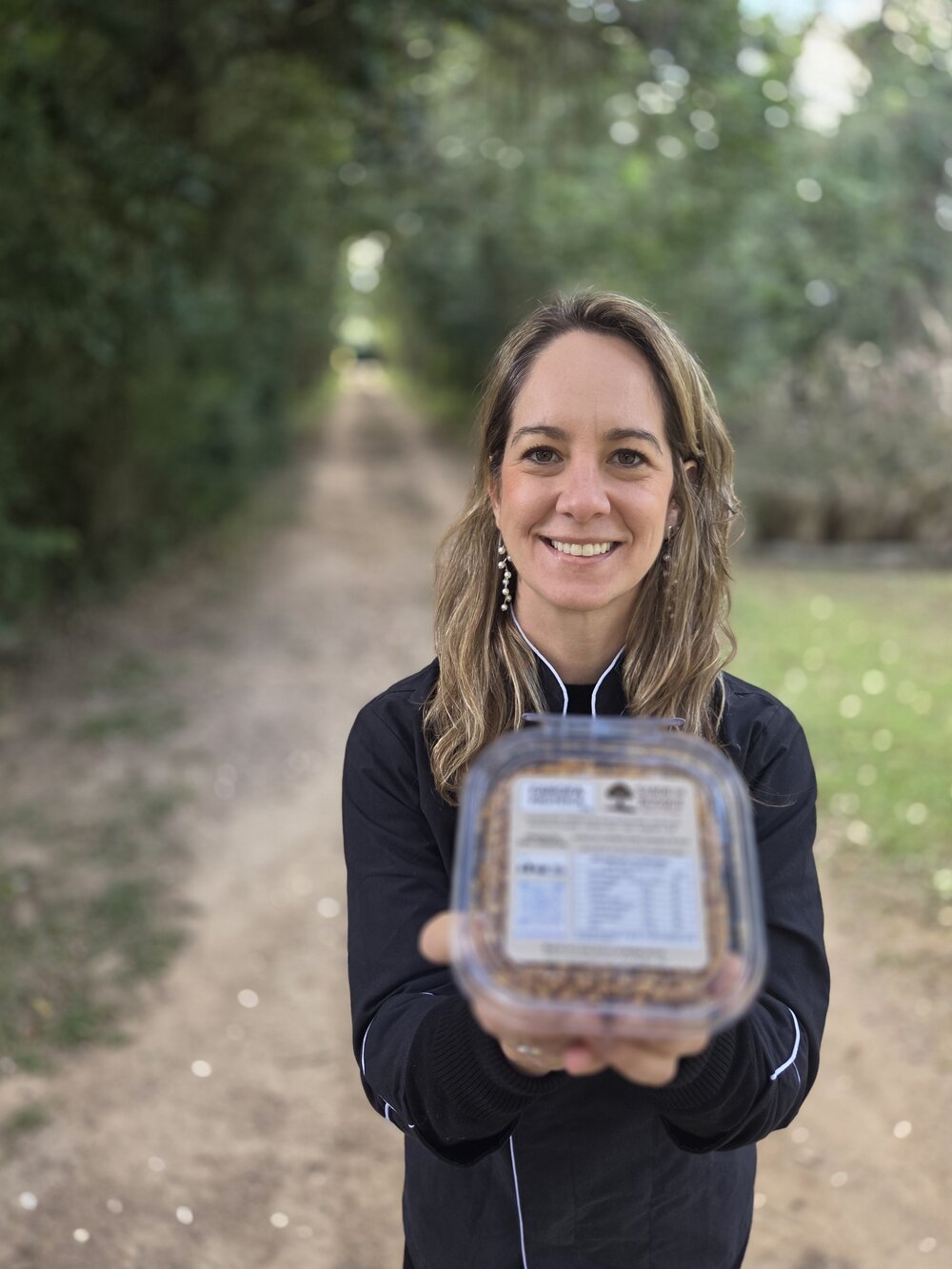 A produtora da Sabor do Bosque segurando um pote de farofa proteica no caminho de árvores
